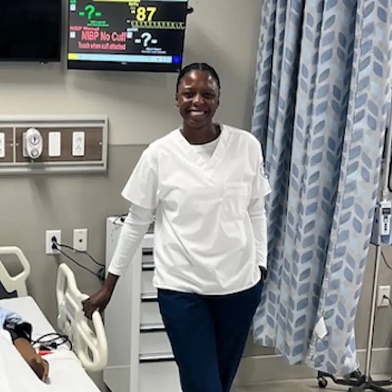 A smiling Khadijah Faison in scrubs stands confidently in a hospital room. The background features medical equipment, including a monitor displaying vital signs, and a hospital bed. The individual exudes a sense of warmth and professionalism, ready to assist patients.