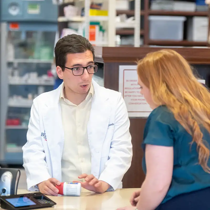 A male pharmacist in a white lab coat is speaking with a female customer at a pharmacy counter. The pharmacist holds a prescription bottle, gesturing while explaining something. The customer, with long red hair, appears attentive. In the background, there are shelves stocked with various pharmacy items.