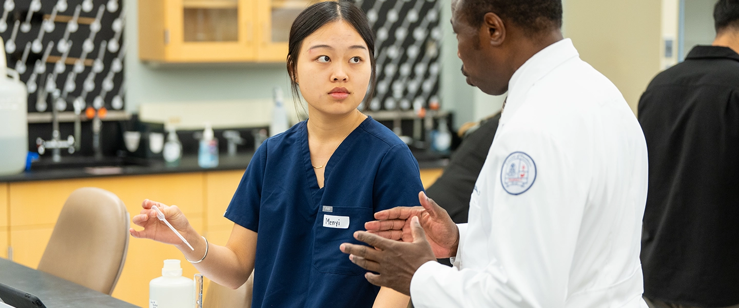 Belmont Doctor of Pharmacy student in navy scrubs uses a pipette during a lab exercise while speaking with a faculty member in a white coat in a campus laboratory.