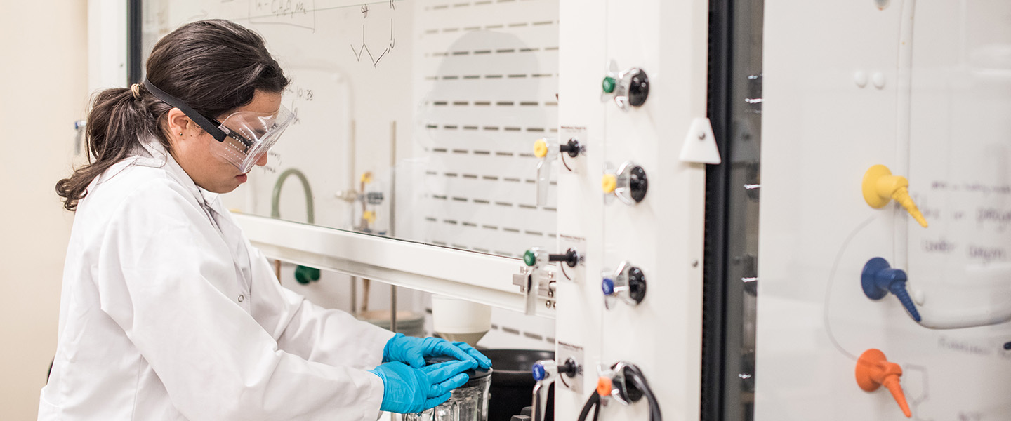 A female student dressed in a white lab coat and goggles prepares a blender.