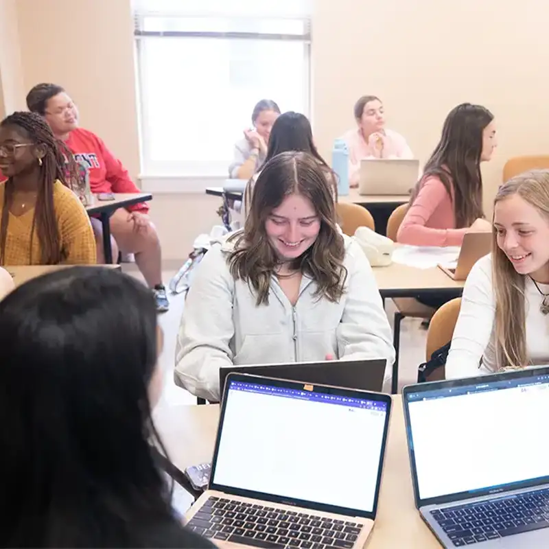 A classroom scene featuring a diverse group of students engaged in collaborative work. Some students are seated at desks using laptops, while others appear to be in discussion. Two students in the foreground share a noticeable moment of laughter and concentration as they look at their screens. Natural light streams in from a window, creating a warm and inviting atmosphere.