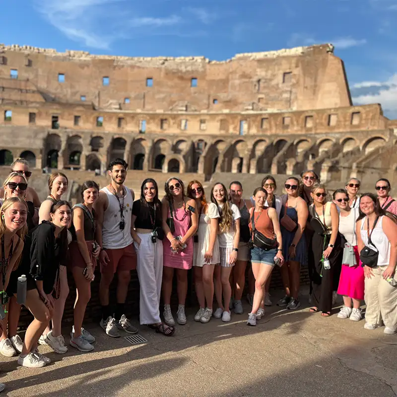 A large group of smiling young adults posing together for a photo inside the Colosseum in Rome, Italy, on a sunny day. The ancient stone arches and walls of the historic amphitheater form the backdrop. Most people in the group are casually dressed and wearing sunglasses.