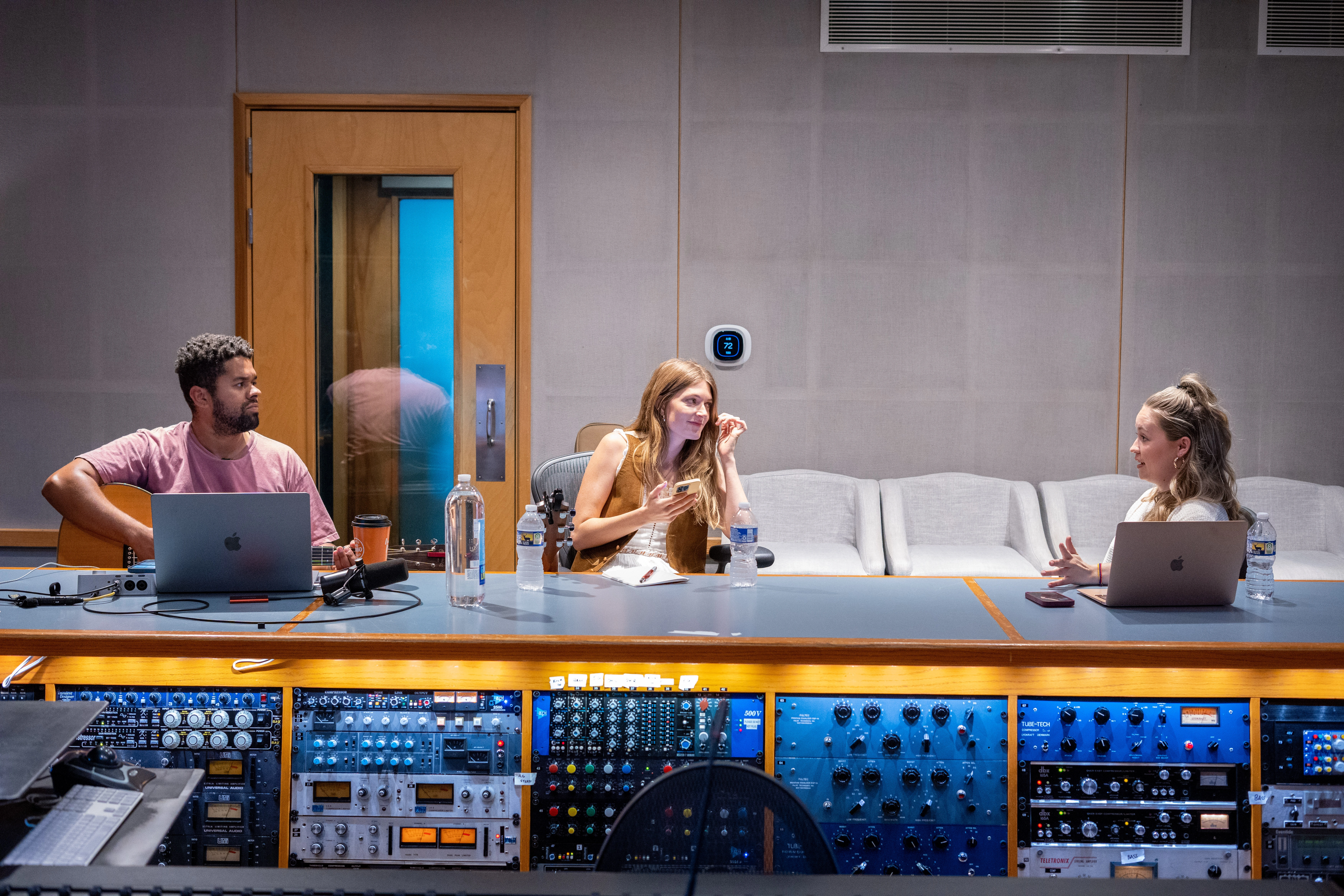 Three people in a recording studio discuss around a mixing console. Laptops open in front of them indicate a collaborative, creative session.