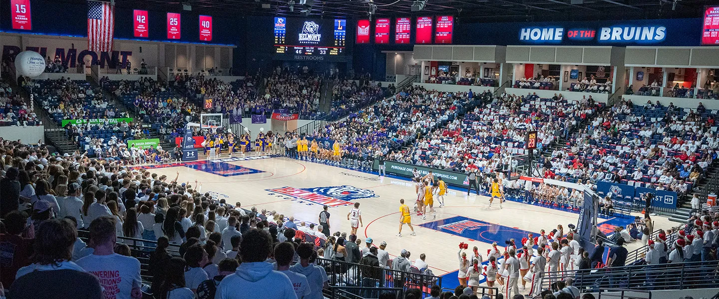 Wide-angle view of the Curb Event Center during a basketball game. The court is surrounded by a large, energetic crowd, with Belmont University's 'Home of the Bruins' banner displayed prominently above.