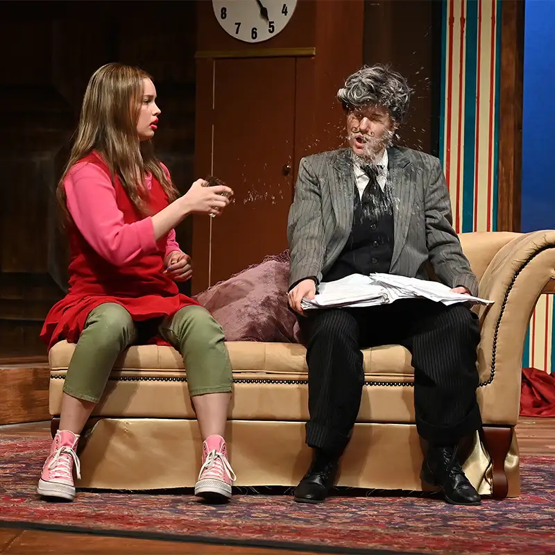 A scene from a theatrical performance featuring a young woman in a red sweater and green pants, sitting on a couch and throwing something at an older man in a suit with a surprised expression, as water splashes from his face.