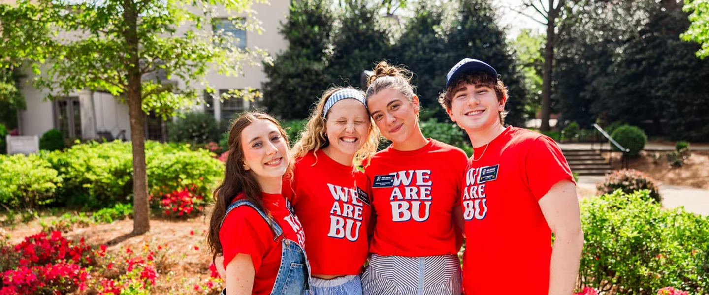Four smiling young people pose together in a sunny garden, wearing matching red “We Are BU” shirts; flowers and greenery create a cheerful campus scene.