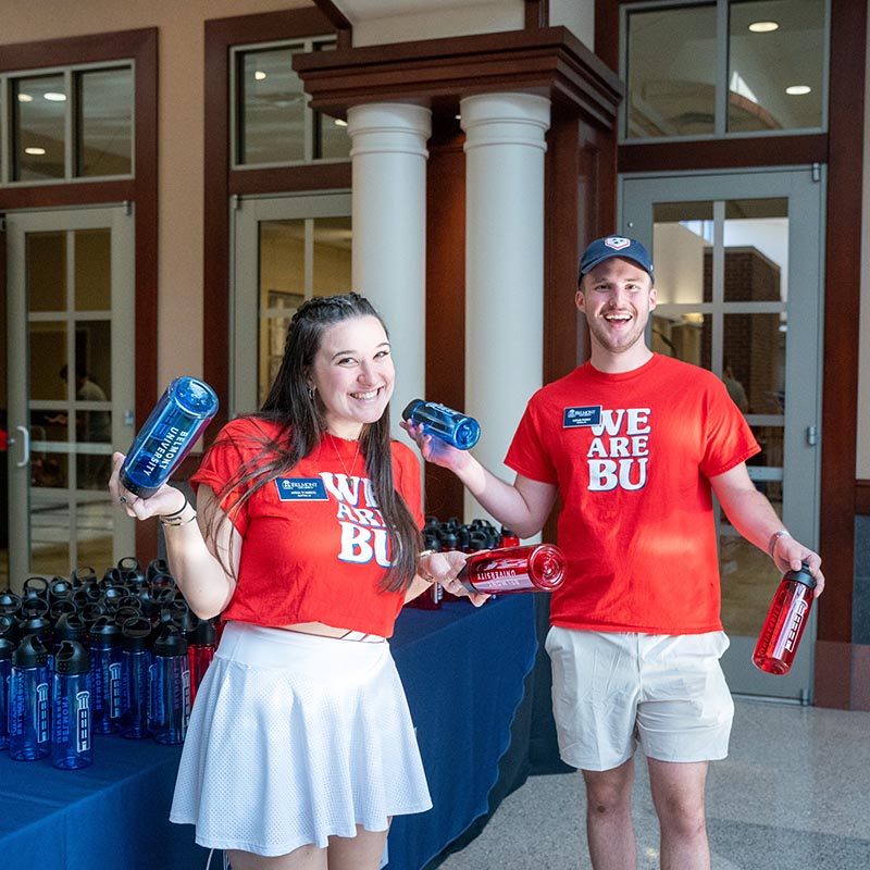 Two student volunteers wearing matching t-shirts smile for camera while setting up Belmont water bottles on a table