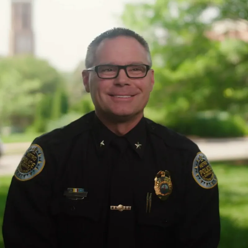 Portrait of Jason Proctor in police captain uniform with glasses, smiling outdoors with green landscaping in the background. 