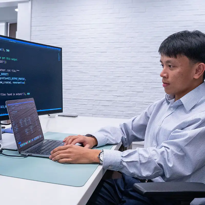 A person sits at a desk working on a laptop connected to a large monitor displaying lines of computer code.