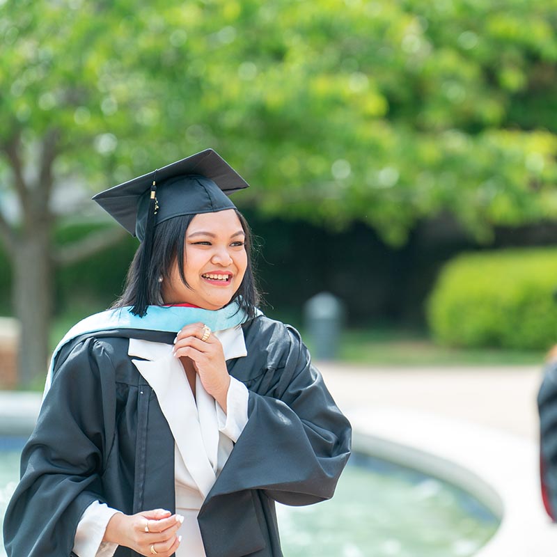 Female student smiling in front of Freedom plaza fountain wearing here master's hood and cap and gown 