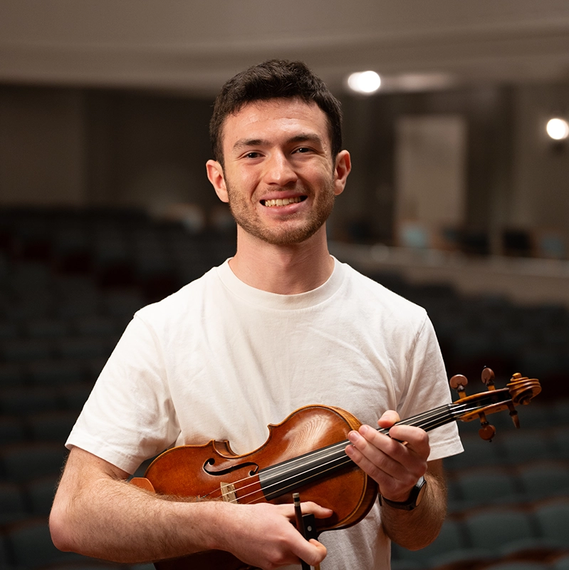 A smiling young musician in a white T-shirt holds a violin and bow while standing in an empty auditorium with rows of seats behind him.