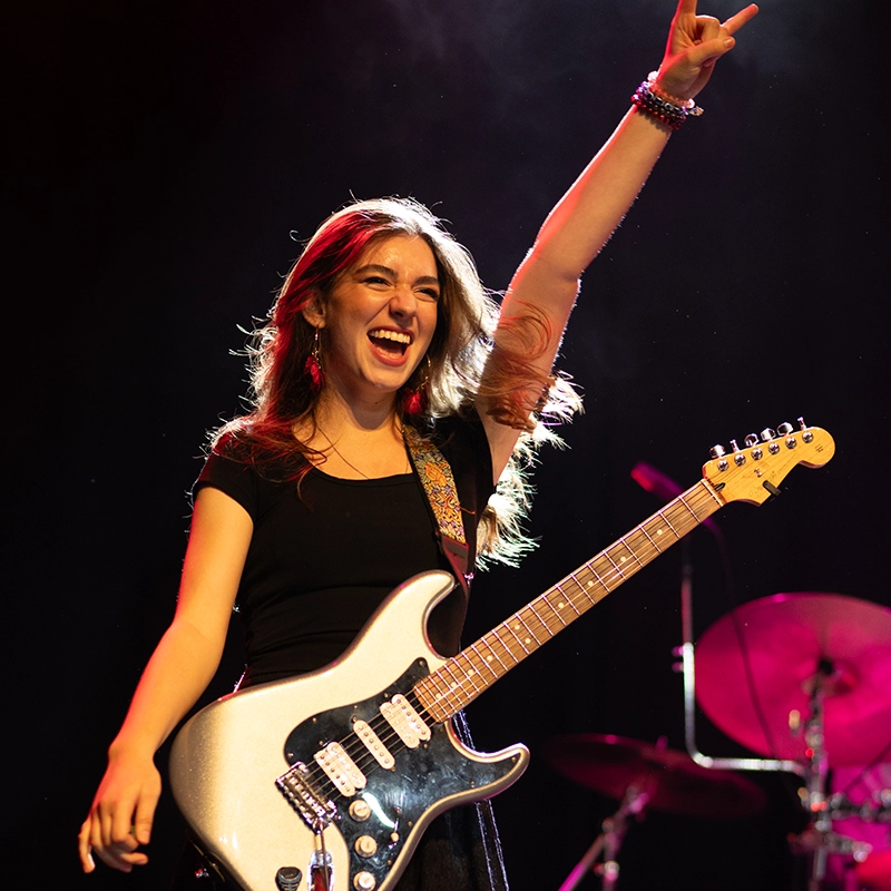 A smiling young woman performing onstage raises one arm triumphantly while holding an electric guitar, illuminated by colorful concert lighting with drums visible in the background.