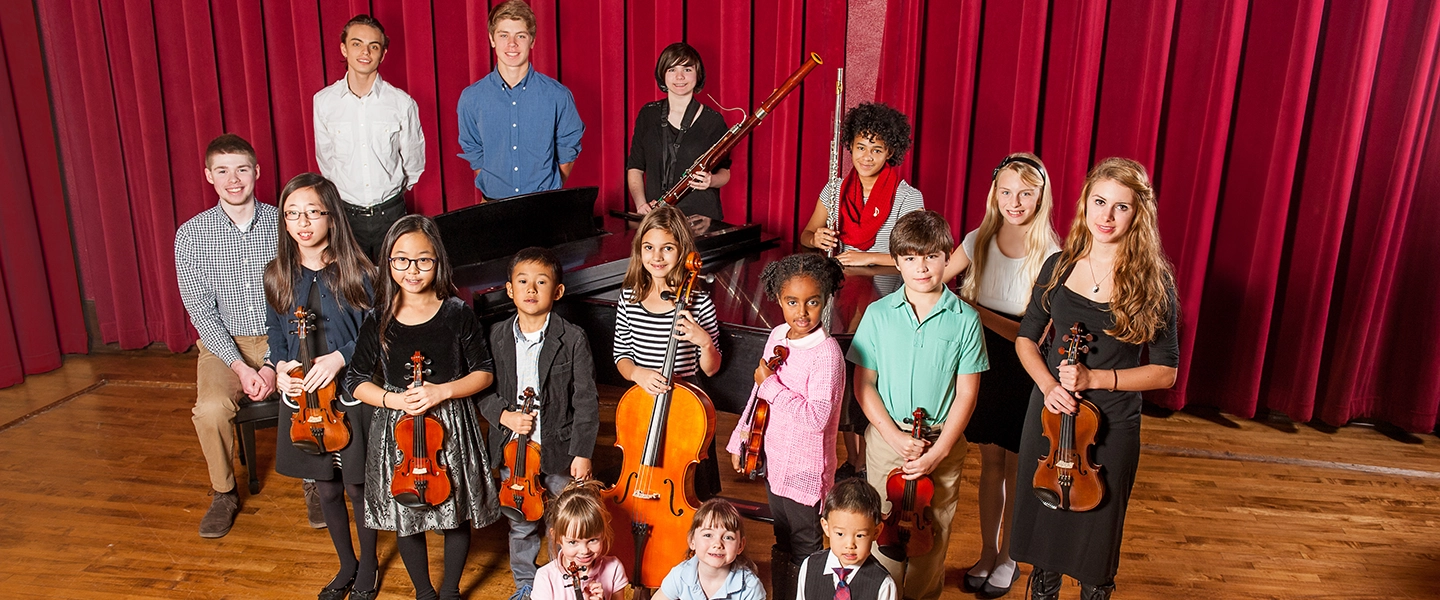 Group portrait of children and teens holding violins, cello, flute and bassoon, standing on a stage in front of red curtains with a grand piano behind them.