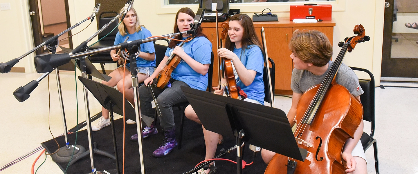A small group of young musicians sit in a recording studio wearing matching blue shirts, holding string instruments including violins and a cello, with microphones and music stands set up around them.