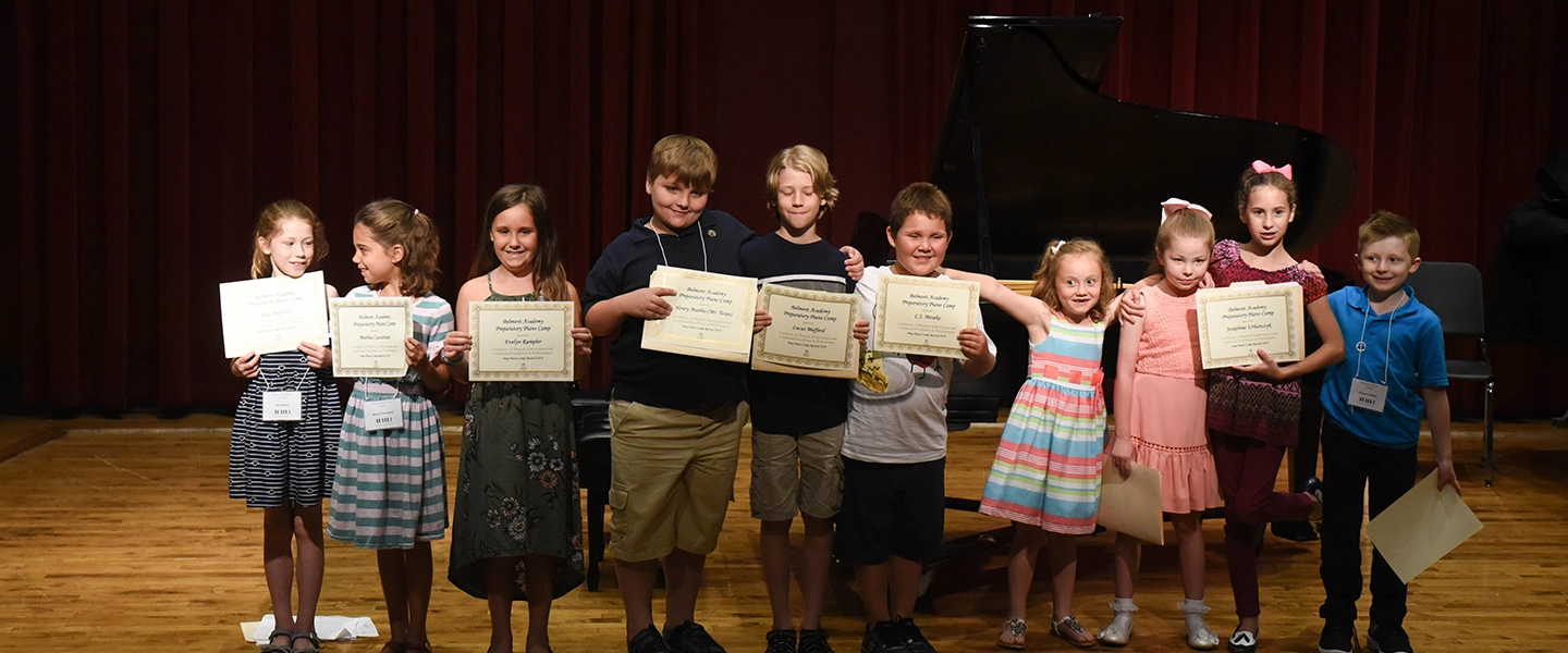 A group of young children stand shoulder to shoulder on a recital hall stage, smiling and holding certificates of completion, with a grand piano and red stage curtains behind them.