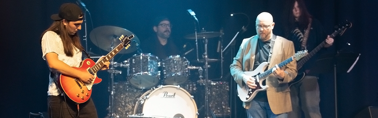 Two guitarists perform onstage under blue stage lighting while a drummer plays behind them, creating a live band performance atmosphere.