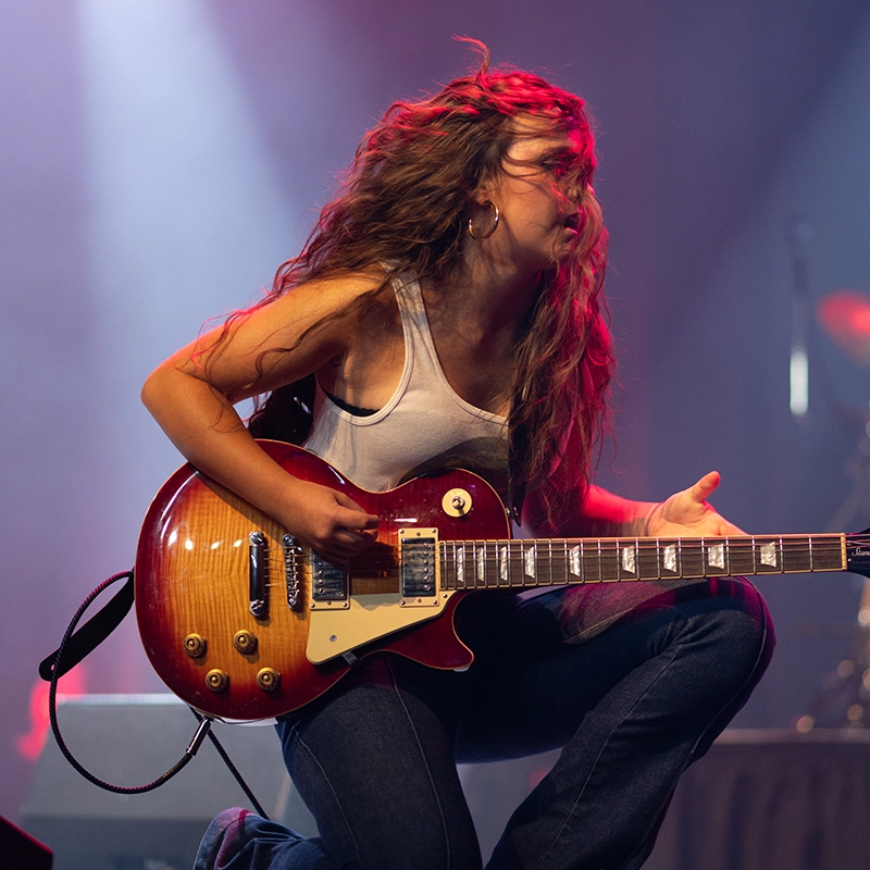 A guitarist with long wavy hair kneels onstage while playing an electric guitar under colorful concert lights, her hair blowing as she performs with high energy.