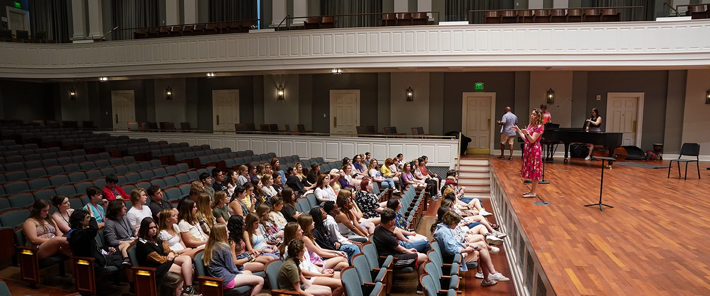 A large group of students sits in an auditorium listening to a woman in a bright pink dress speaking from the stage, with a pianist and several staff members positioned behind her.