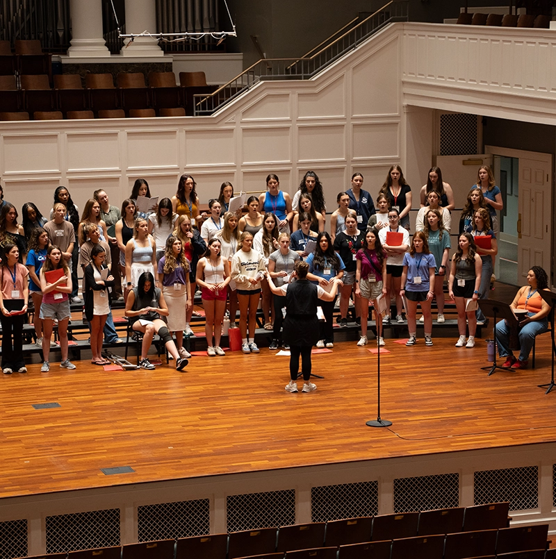A large choir of young singers stands on risers in a concert hall as a conductor leads rehearsal from the front, with a pianist seated to the side.