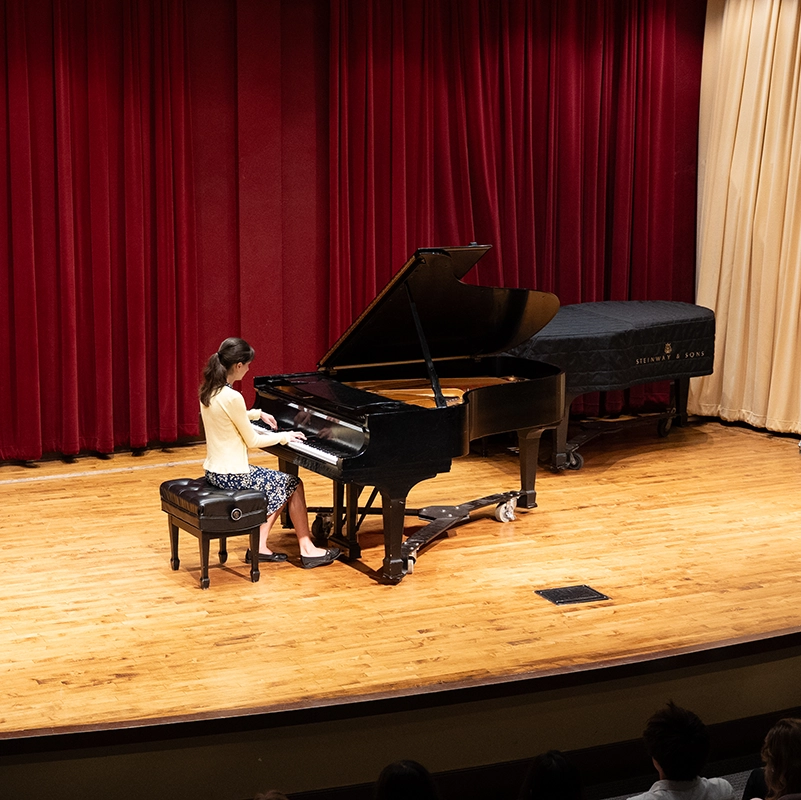 A pianist performs solo on a grand piano on a wooden stage, framed by deep red curtains, while an audience watches from the foreground.