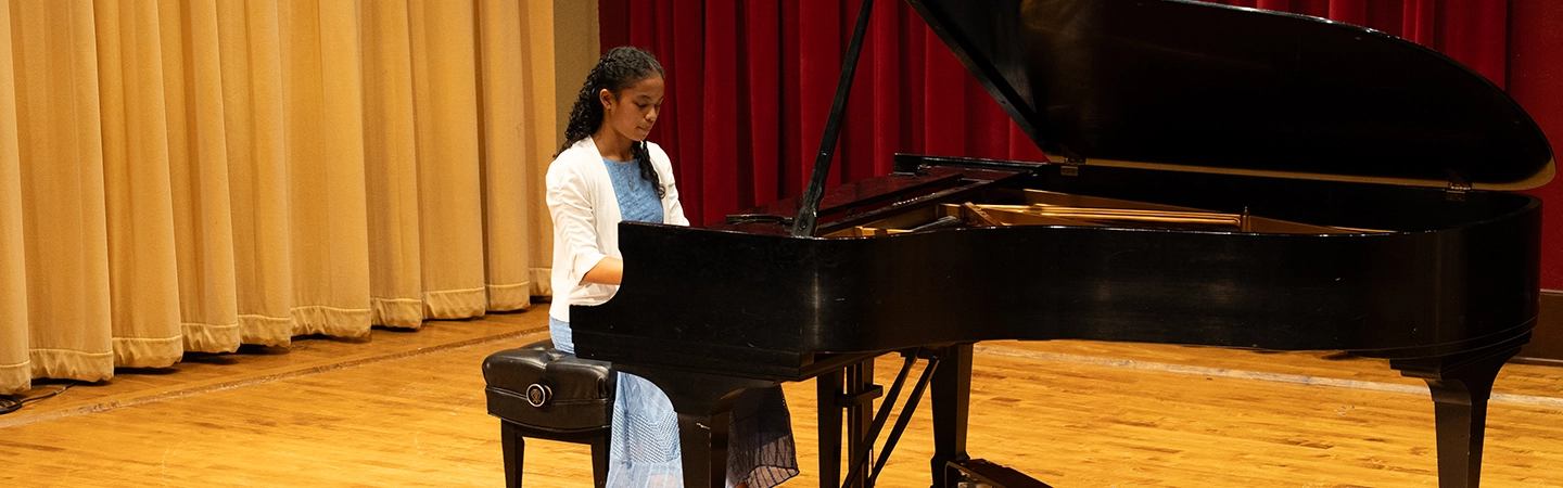 A young pianist in a light blue dress and white cardigan performs on a grand piano on a wooden stage, framed by beige and red stage curtains.