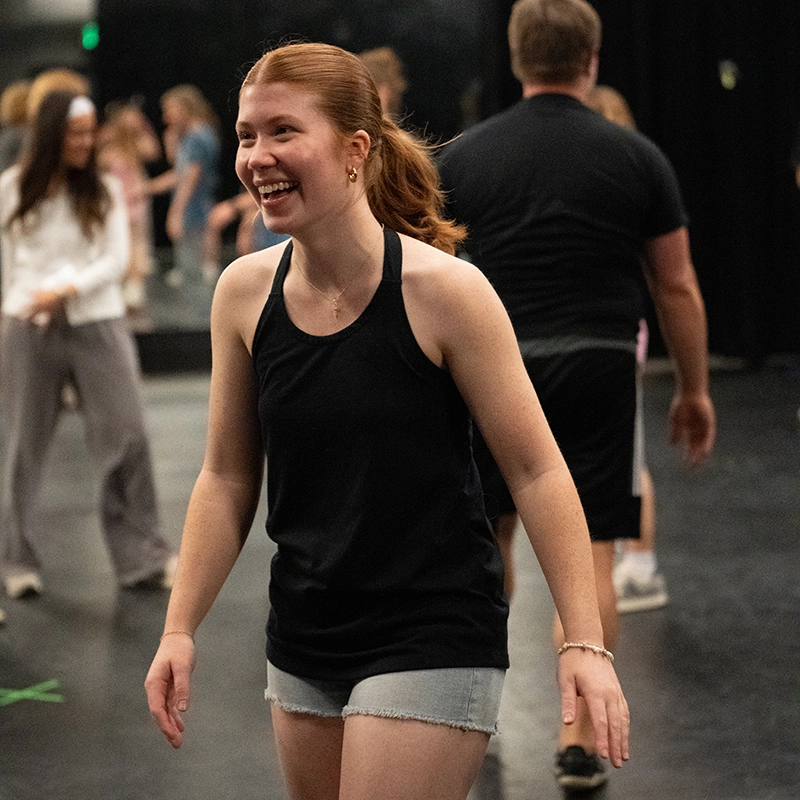 A smiling student in a black tank top and denim shorts participates in a lively dance or movement class, surrounded by other students in a studio setting.