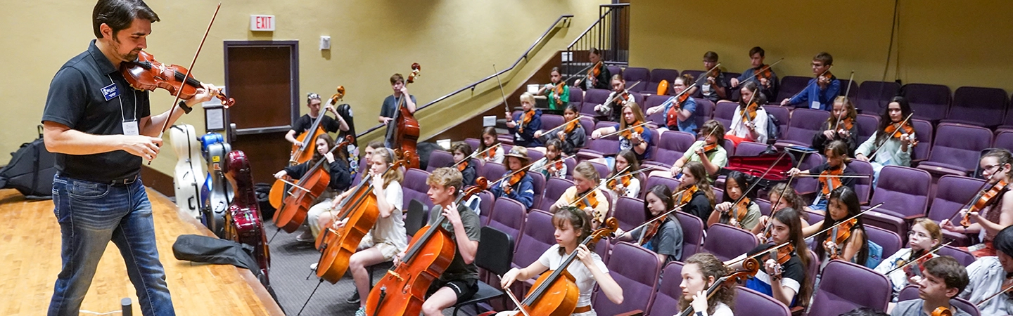 A violin instructor demonstrates onstage while a large group of young string players—holding violins, violas, cellos, and basses—practice together in a tiered auditorium.