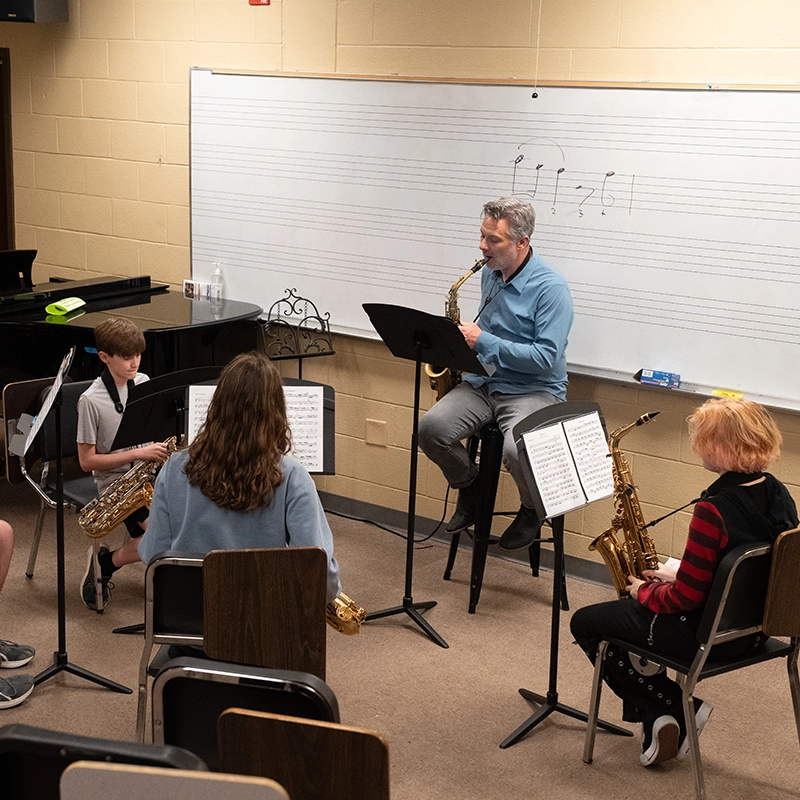 A music instructor plays a saxophone while seated at the front of a classroom, demonstrating for a small group of students holding saxophones. The students sit in a semicircle with music stands, and a whiteboard with musical notation fills the background.