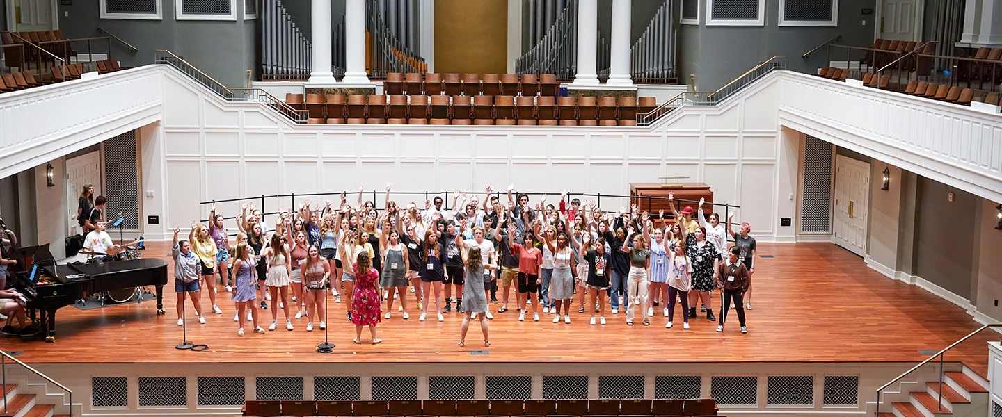A large group of students stands onstage in a concert hall with their hands raised during a choral rehearsal, as instructors lead from the front and a pianist accompanies from the side.