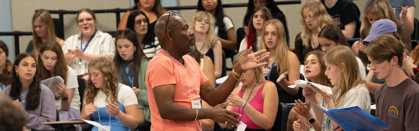 A vocal instructor leads a large group of students in a choral rehearsal, clapping rhythmically as the students follow along with their music folders.