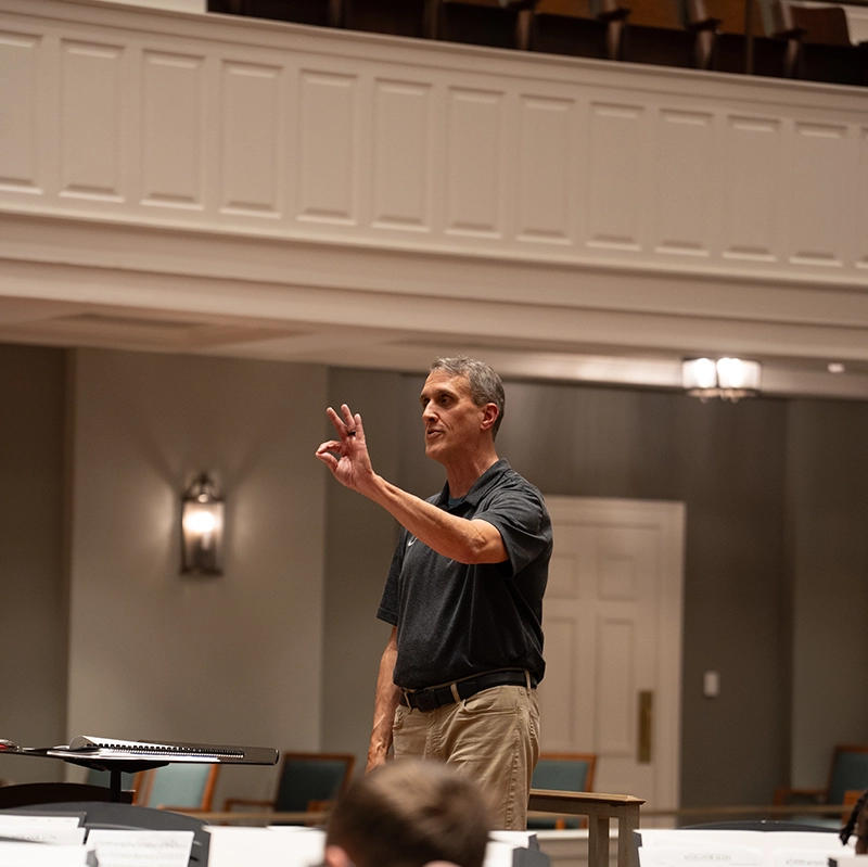 A conductor stands at the front of a rehearsal room, raising his hand to cue musicians, with sheet music and orchestra members visible in the foreground.