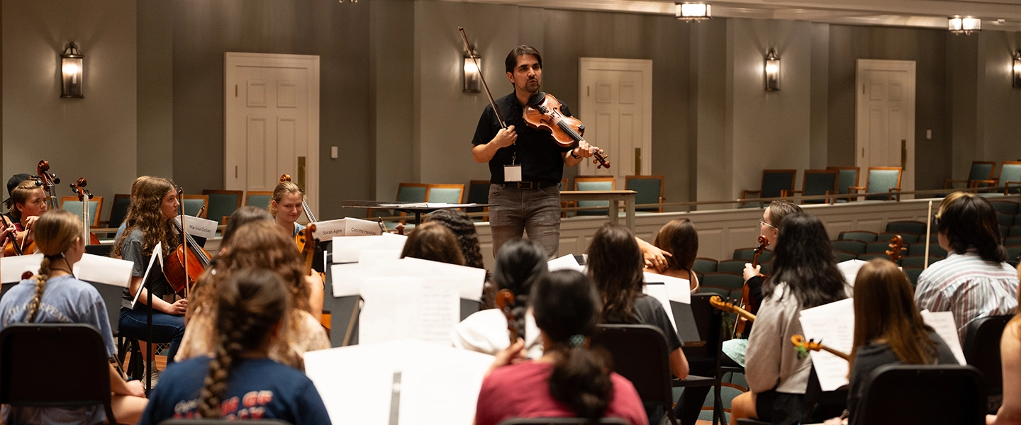 A violin instructor stands at the front of a rehearsal room holding a violin and bow, addressing a large group of young string musicians seated with their instruments and sheet music.