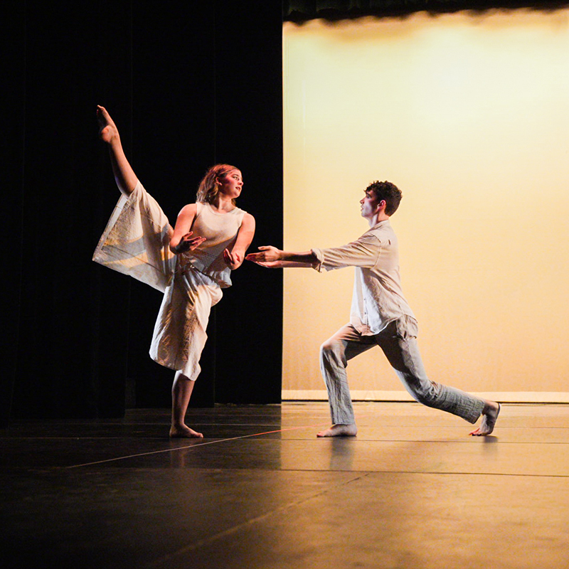 Two dancers perform on stage, one balancing with a raised leg while the other lunges forward with outstretched arms under warm, minimal stage lighting.