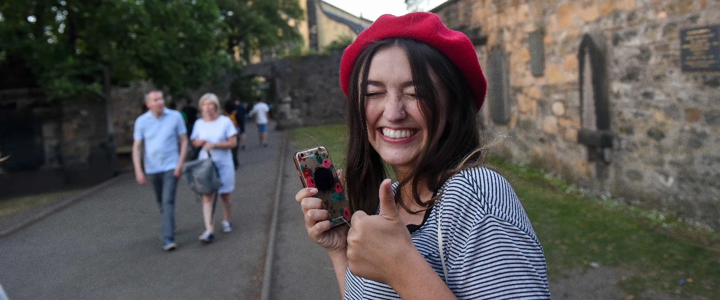 Happy young woman in red beret gives thumbs up, holding phone, with a stone building backdrop.