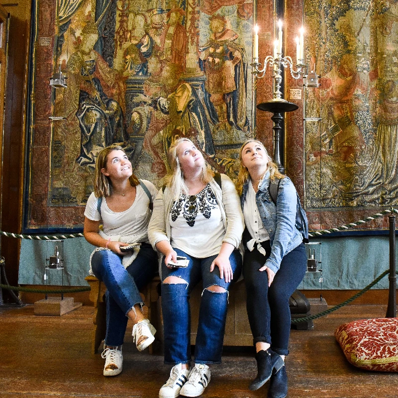 Three women admire a detailed historical tapestry and candelabra in a museum, looking upwards.