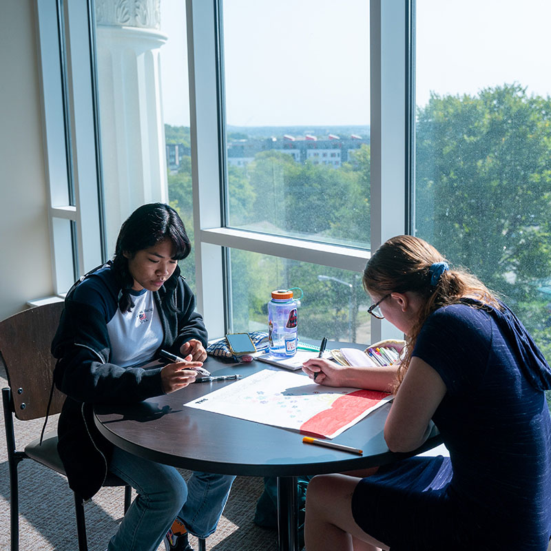 Two students working together at a table next to a window with a view of 15th ave