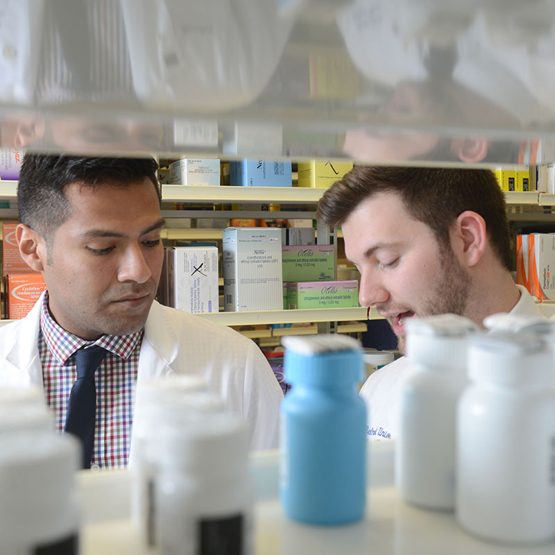 A view of two Pharmacy students discussing a medication from the other side of a medicine shelf