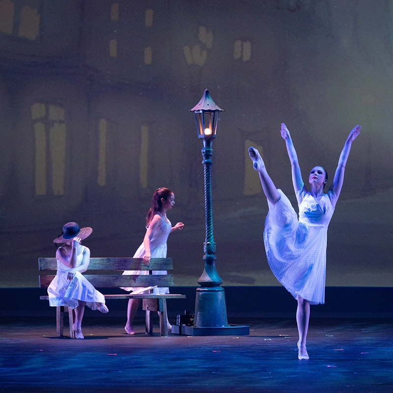 Three ballet dancers perform on stage in an elegant scene. One dancer is posed with a raised leg while the other two sit on a bench, all wearing white dresses.