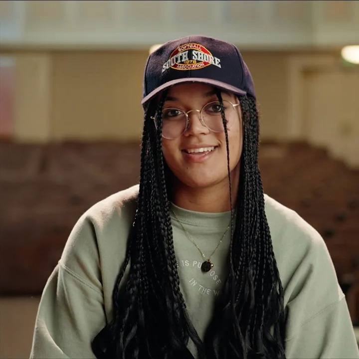 Rue Bradfield wearing glasses, and a baseball cap sits in an auditorium, wearing a sage green sweatshirt with a relaxed, welcoming expression.