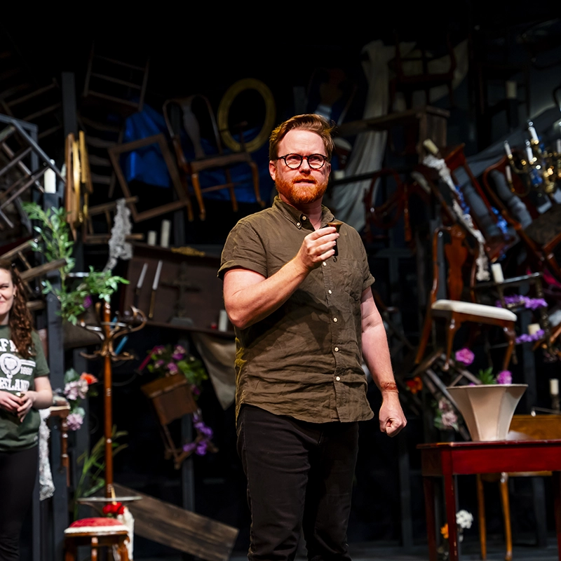 A man with a red beard and glasses performs on stage during a theater production, speaking passionately into a microphone, with a woman visible behind him.