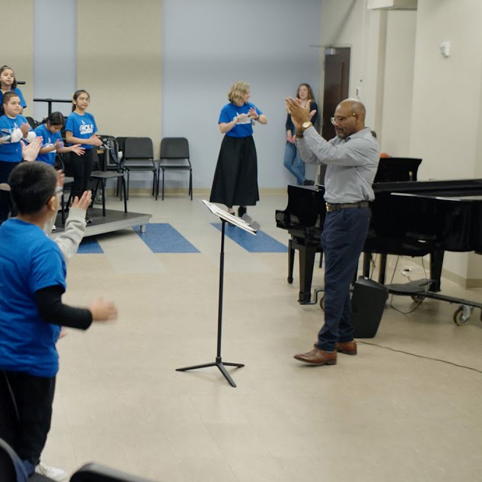 Instructor claps while leading a group of young students in blue shirts during choir rehearsal in a classroom with risers, piano, and music stands.