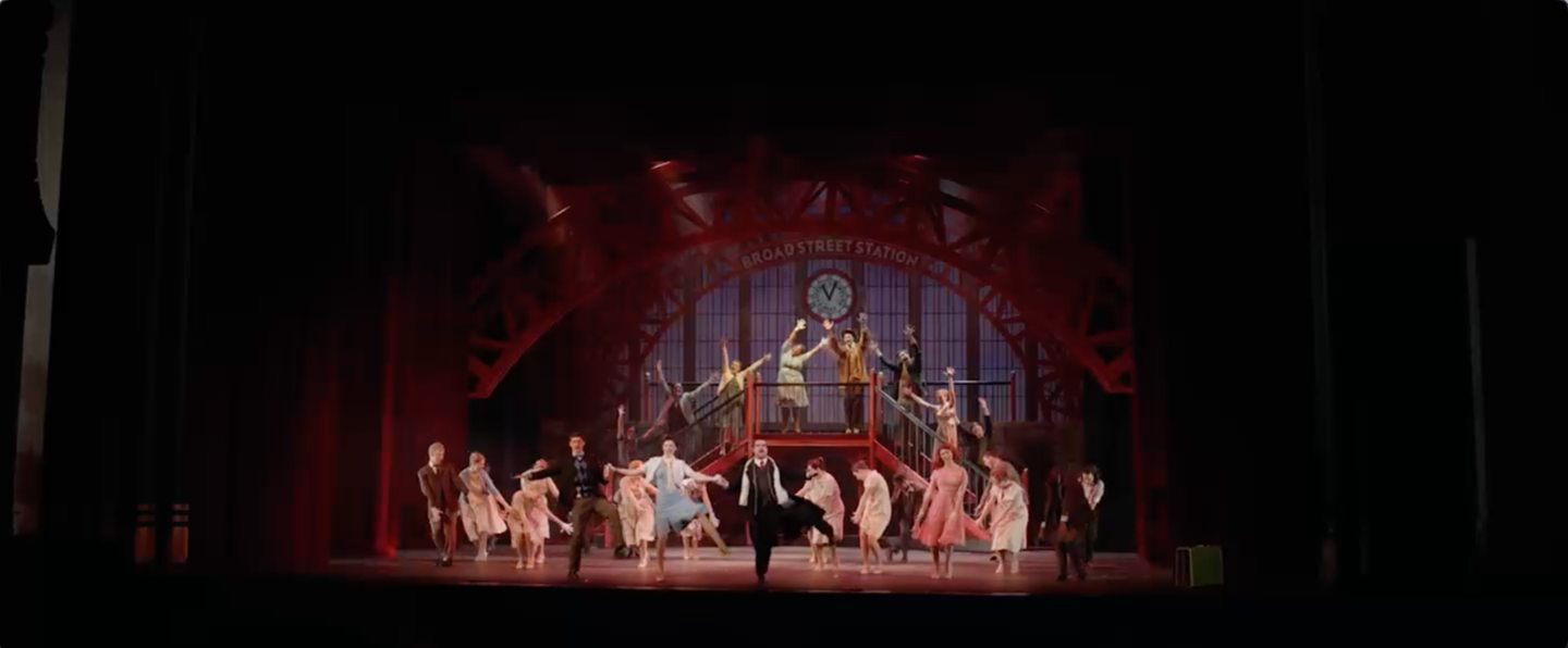 A large group of performers dance energetically on a theater stage set resembling a train station, with a staircase, clock, and red metal arch in the background.