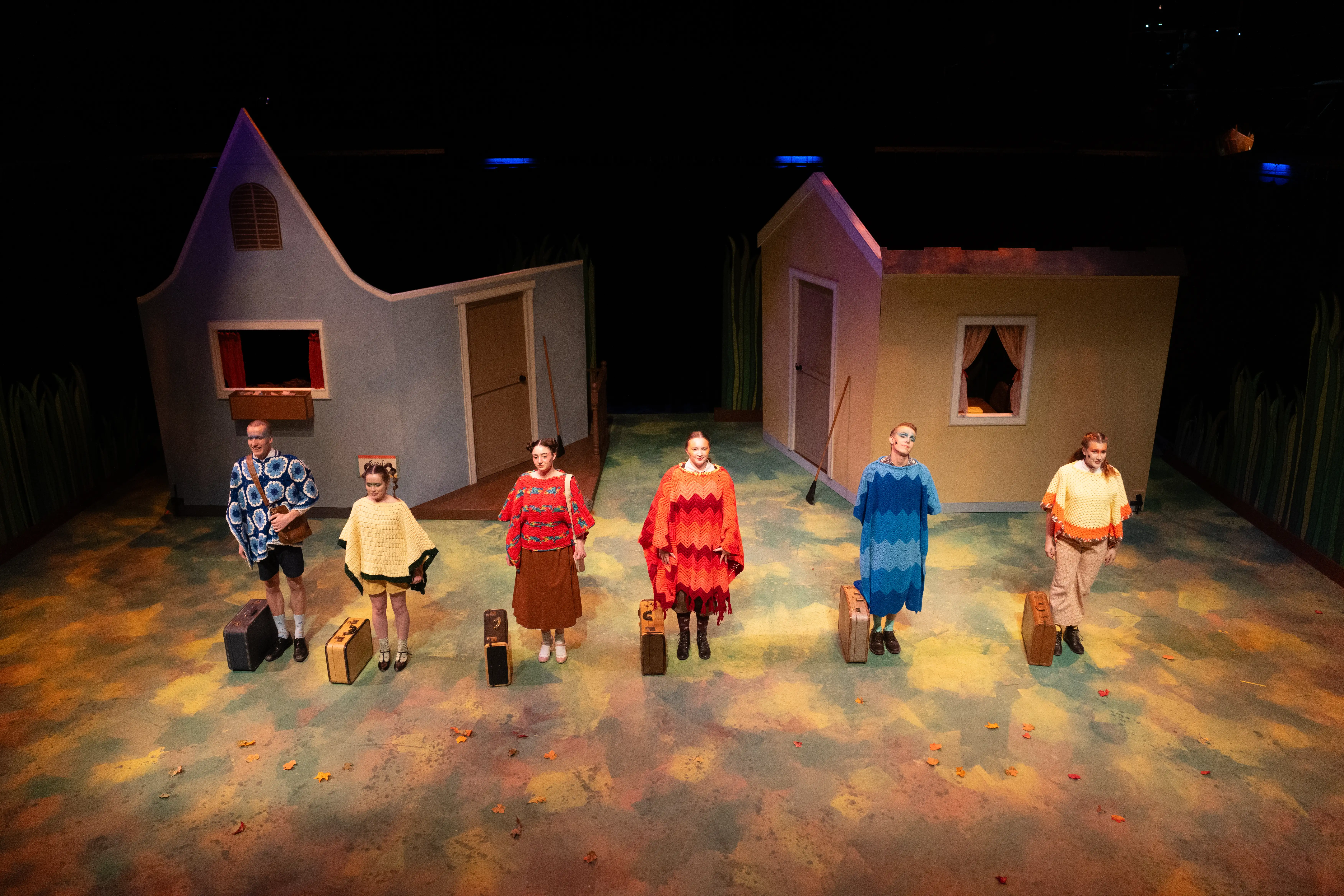 Actors in colorful ponchos stand in a line holding suitcases on a whimsical set with cartoon-style houses during a production at the Black Box Theater.