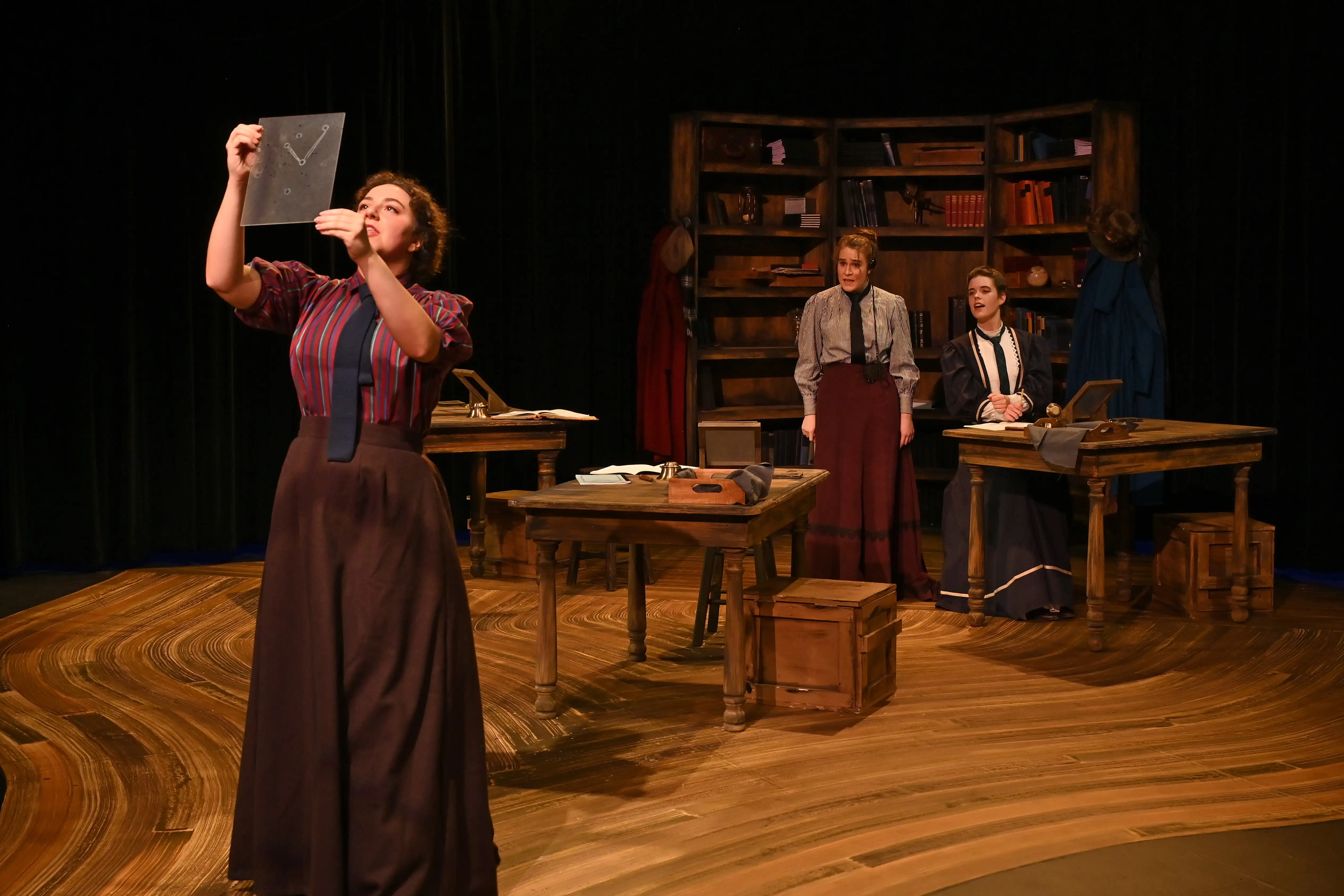 Actors perform in period costumes on a vintage office set in Belmont’s Black Box Theater, with desks, props, and a large bookcase.