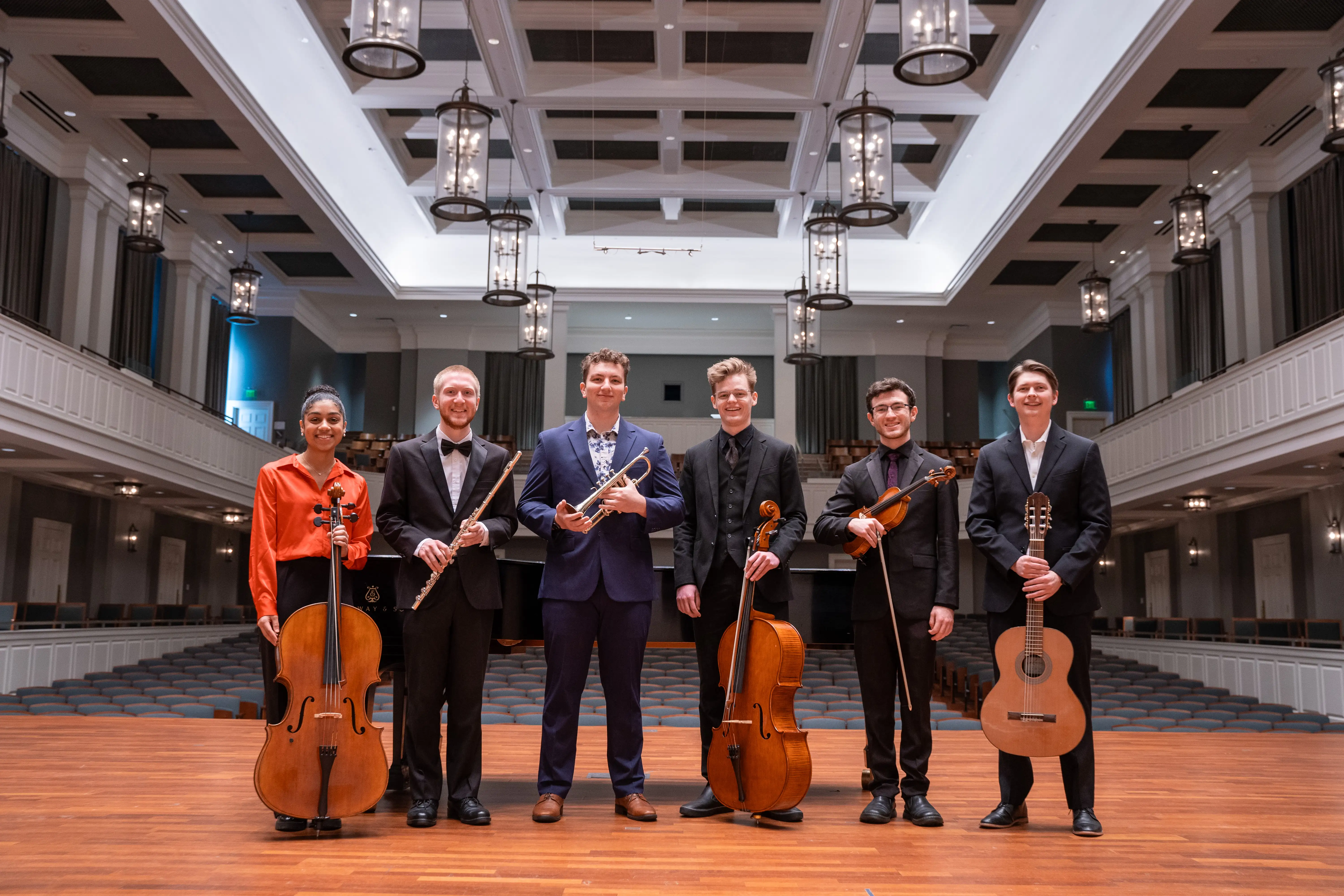 Six student musicians from the Honors Orchestra stand proudly onstage in McAfee Concert Hall holding their instruments, posing for a group portrait under elegant lighting.
