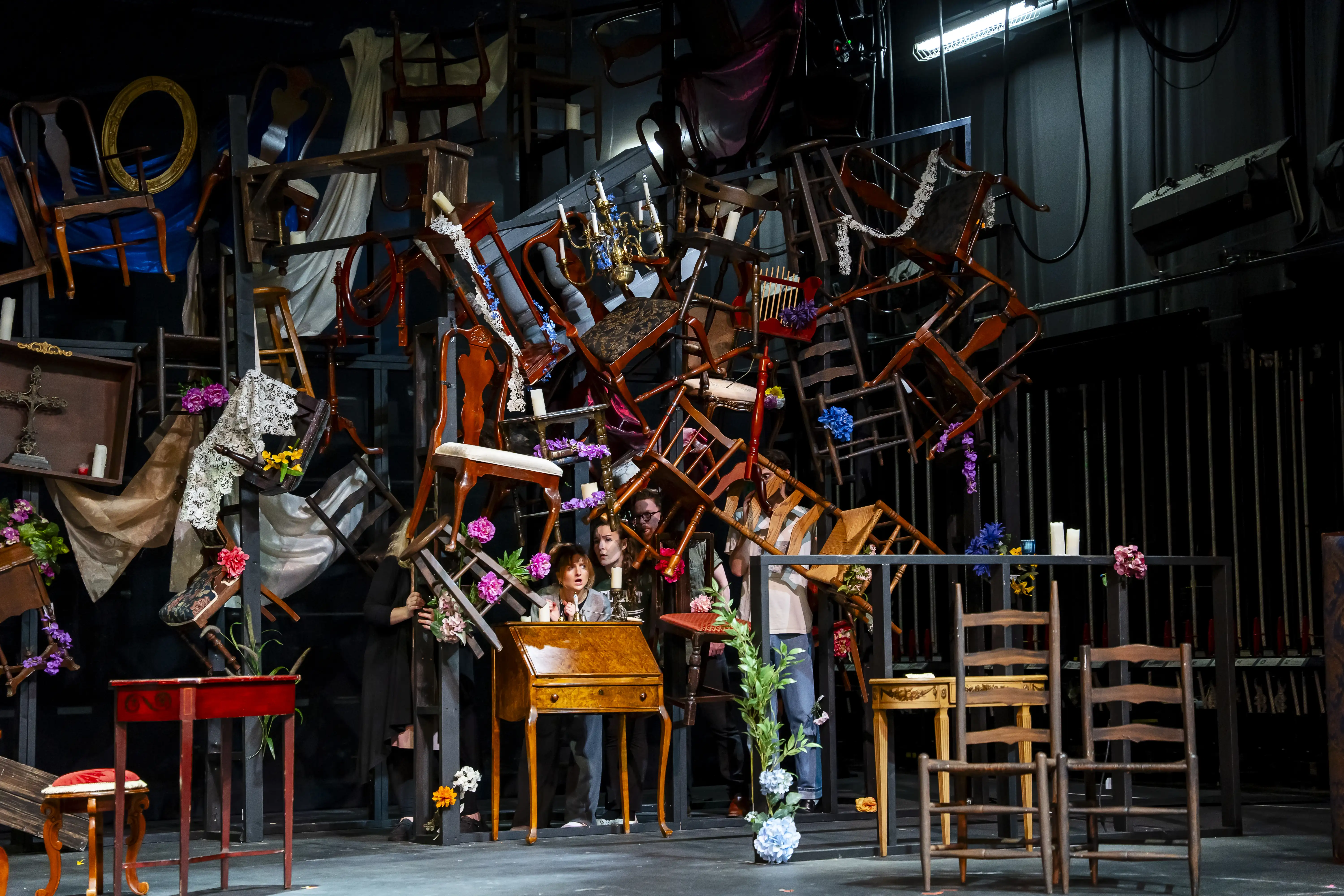 Actors emerge from a chaotic set of chairs, flowers, and props stacked in a dramatic arrangement during an experimental play at Troutt Theater.