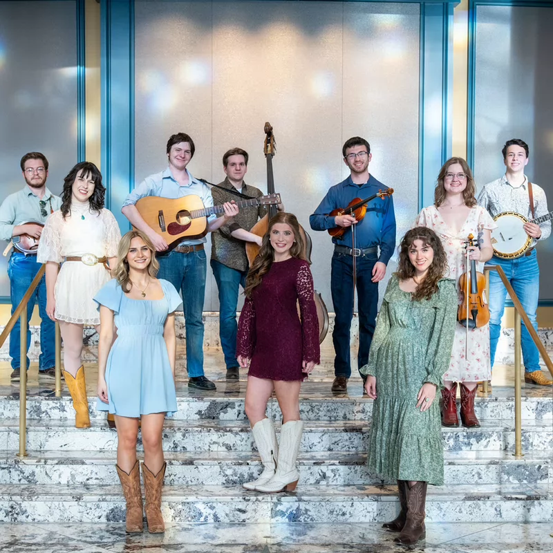 A diverse bluegrass band poses on grand marble steps, holding guitars, banjos, violin, and other instruments in a bright, welcoming hall.