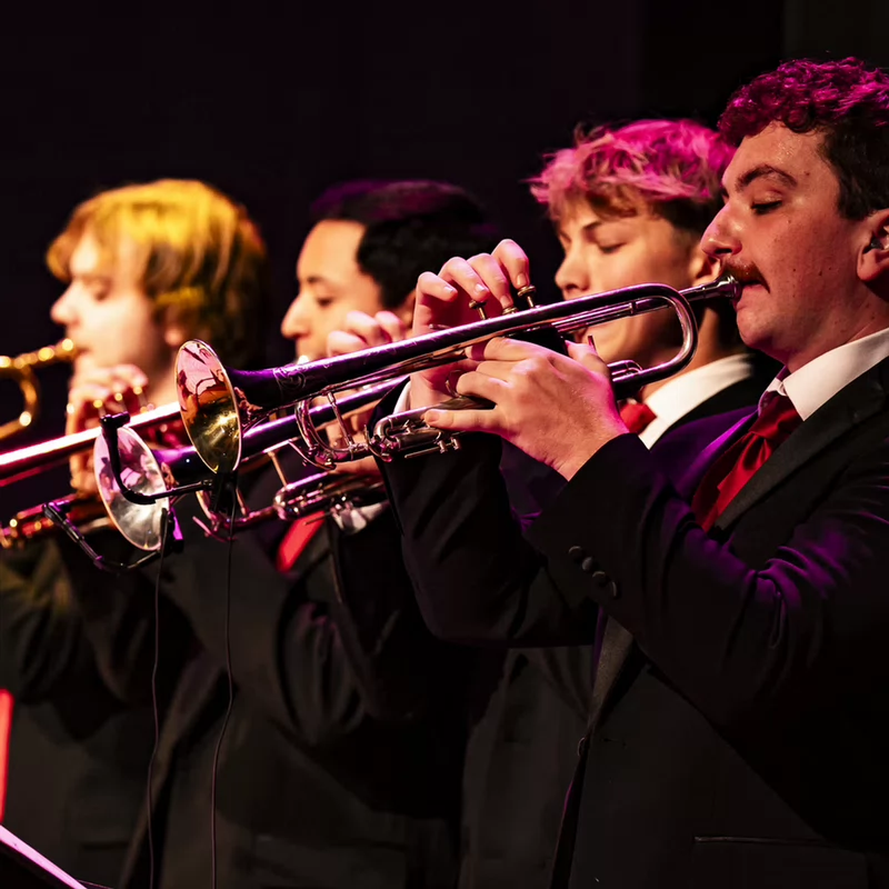 Four trumpet players in suits and ties perform on stage, each holding a trumpet to their lips, while reading sheet music.