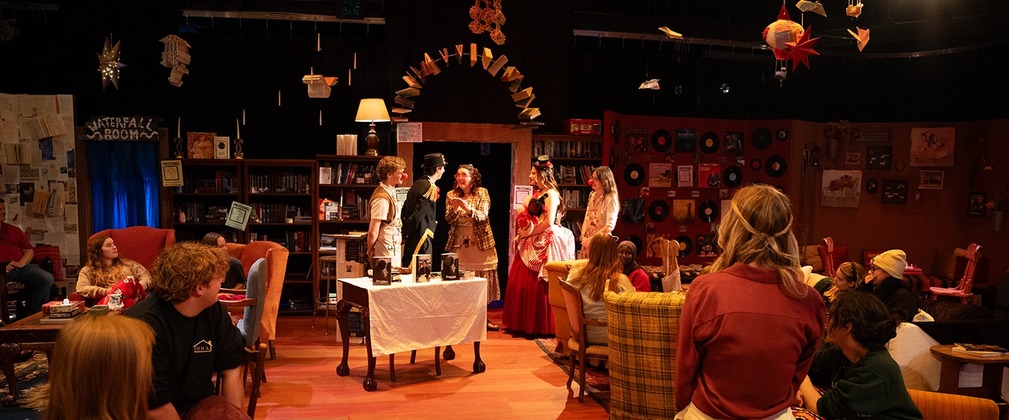 Audience members sit around a staged living-room set as actors perform a theatrical scene under warm lighting, with bookshelves, records and hanging decor on stage.