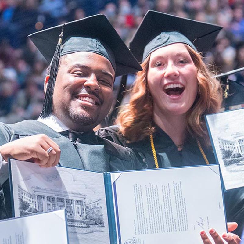 3 students sitting at graduation ceremony in cap and gown as they hold their diplomas and pose for a picture